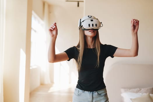 A young woman stands in a bright room wearing a virtual reality headset. She appears immersed in an interactive experience, moving her arms as if engaging with a digital landscape.