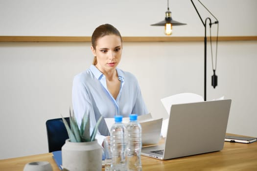 A woman in a light blue blouse is intently reviewing papers at a modern desk. She is surrounded by a laptop, water bottles, and a small plant, all set in a bright office space with minimal decor.