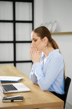 A business woman sits at her desk with a laptop, hands clasped in front of her mouth, visibly deep in thought and reflection