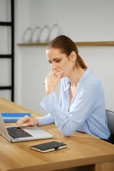 A young woman dressed in a light blue shirt is focused on her laptop while sitting at a wooden desk. She appears deep in thought, resting her chin on her hand in a contemporary office setting.