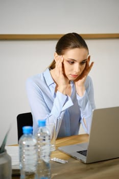 A young woman sits at a wooden desk, visibly stressed while working on her laptop.