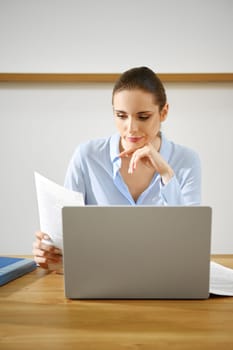 A sceptical woman is engaged in reviewing important documents while simultaneously working on her laptop at a tidy wooden desk