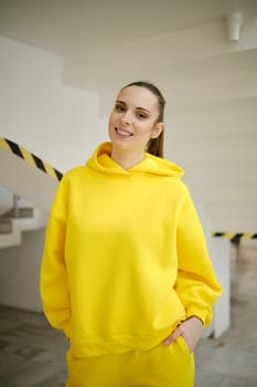 A woman wearing a vibrant yellow hoodie smiles warmly while standing in a contemporary building.