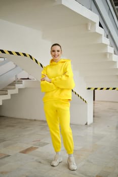 A woman stands on a staircase wearing a bright yellow hoodie and matching pants. She smiles confidently against a backdrop of clean lines and contemporary design, creating a striking visual.