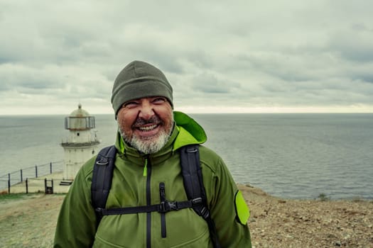 A man stands by the sea, wearing a green jacket and hat. He smiles broadly while enjoying the coastal view. A lighthouse is visible in the background, surrounded by grey clouds.