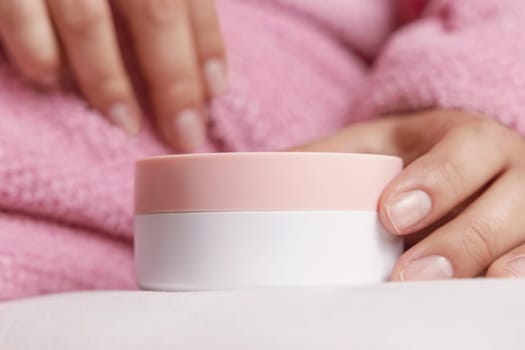 Woman in pink bathrobe puts cream jar on white bedsheet closeup, cosmetic mockup. Organic products for body and face care, beauty and spa procedures. Girlish bodycare concept