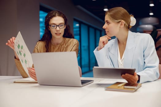 Two businesswomen discuss innovative ideas in a modern office while reviewing information on a laptop