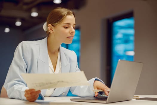 A dedicated and focused professional meticulously reviewing various documents on her laptop in a modern office setting