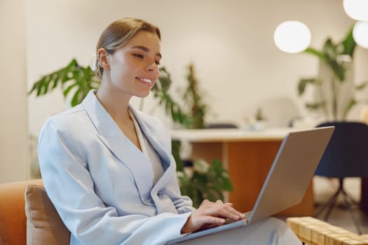 A focused woman dressed in a stylish light suit is deeply engaged with her laptop in a chic office setting