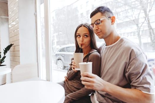 A young couple sits closely together in a bright cafe, sipping from to-go cups. Natural light fills the space, enhancing the warm atmosphere of their shared moment.