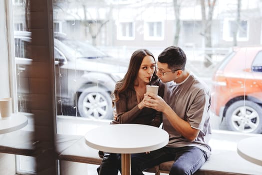 Inside a cozy cafe, a couple shares a drink while sitting close together. Snowfall is visible outside, creating a romantic winter atmosphere.