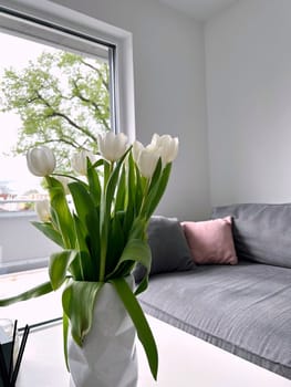 Fresh white tulips arranged in a sleek white vase, positioned on a stylish white table next to a soft gray sofa, natural light streaming through large window, creating a serene and inviting atmosphere