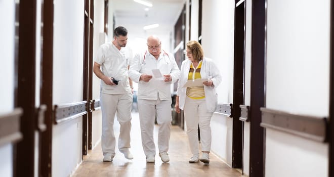 A group of healthcare professionals in white uniforms walking through a hospital hallway while reviewing patient documents and discussing treatment plans