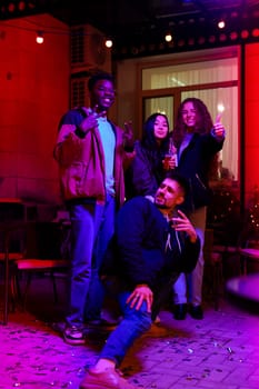Four friends are joyfully posing with drinks and sparklers at a vibrant outdoor venue decorated with colorful lights during a nighttime celebration.