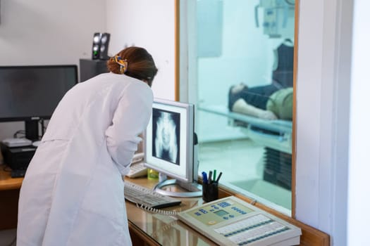 A radiologist in a white lab coat sitting at a workstation while analyzing an X-ray image of a patients hip in a medical imaging room.
