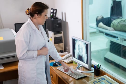 A radiologist in a white lab coat sitting at a workstation while analyzing an X-ray image of a patients hip in a medical imaging room.