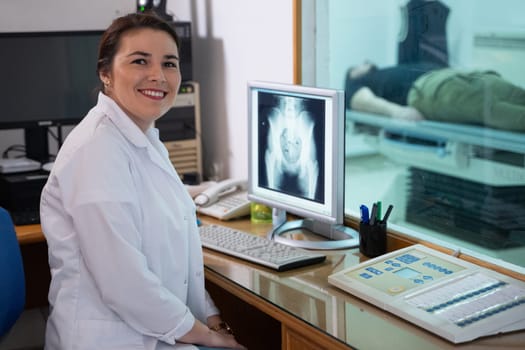 A radiologist in a white lab coat sitting at a workstation while analyzing an X-ray image of a patients hip in a medical imaging room.