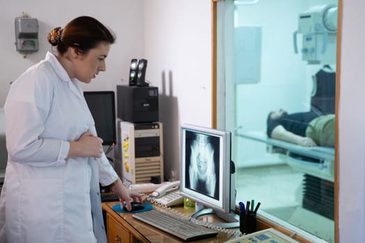 A radiologist in a white lab coat sitting at a workstation while analyzing an X ray image of a patients hip in a medical imaging room