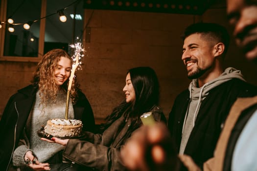 A group of friends joyfully celebrates a birthday at night. One friend holds a beautifully decorated cake with a sparkler, while others share smiles and laughter, enjoying the festive atmosphere.