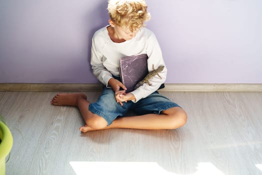 A young boy sits cross-legged on the floor, gently holding a lizard in one hand and a book in the other. Sunlight streams into a softly colored room.