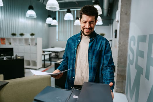 A man stands in a contemporary office, smiling as he examines papers. The space features modern decor, bright lighting, and workstations in the background.