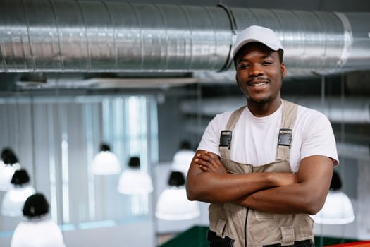 A young man in a casual outfit poses with crossed arms in a contemporary work environment featuring industrial design. The background highlights distinctive light fixtures and exposed ducts.