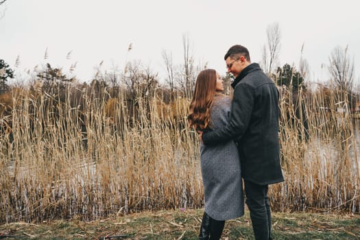 A couple stands closely by a calm waterway, sharing a warm embrace amidst tall reeds. The tranquil autumn setting creates a cozy atmosphere as they enjoy each other's company.