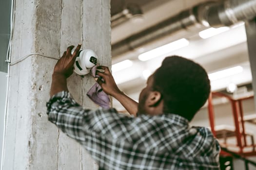 A worker in casual attire is using a cloth to clean a surveillance camera mounted on a concrete pillar, ensuring the equipment functions properly in a modern industrial environment.