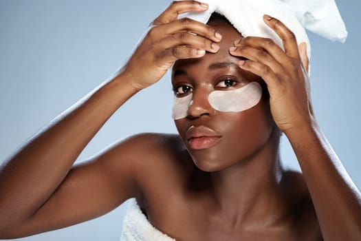 A woman with a towel wrapped around her head gently applies eye patches to her skin, focusing on her beauty routine in a serene indoor setting.