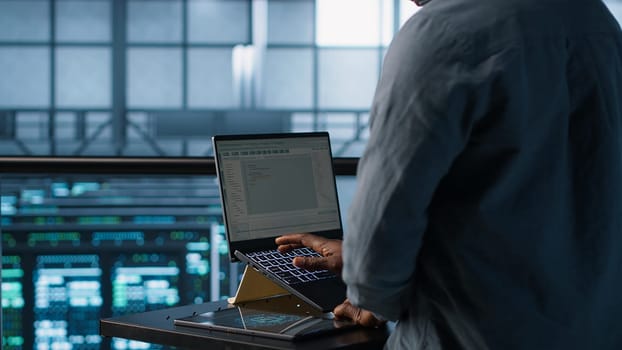 Engineer in data center operating notebook, deploying new server architectures. Server farm employee verifying settings using laptop, maintaining stable network performance, camera A