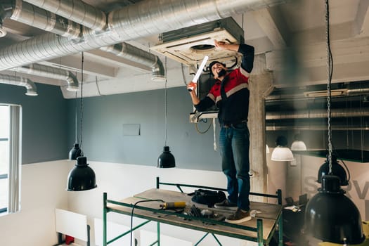 Technician is adjusting an air conditioning unit on the ceiling while standing on a scaffold. Modern office workspace features industrial design with exposed ducts.