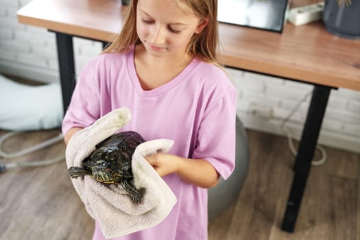 A young girl in a purple shirt gently cradles a turtle wrapped in a towel while standing in a warm, inviting indoor environment filled with natural light.