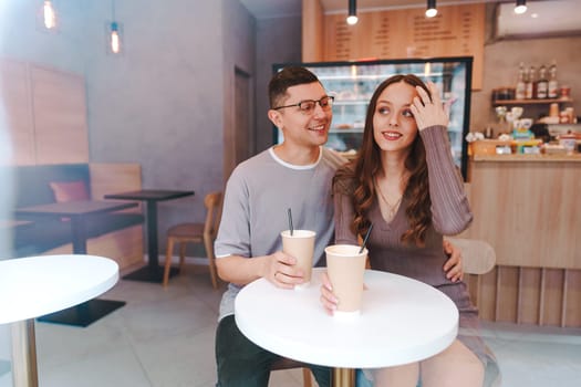 A couple sits close together at a small table in a bright cafe, sipping coffee from paper cups. They are smiling and engaging in a lighthearted conversation, creating a warm atmosphere.
