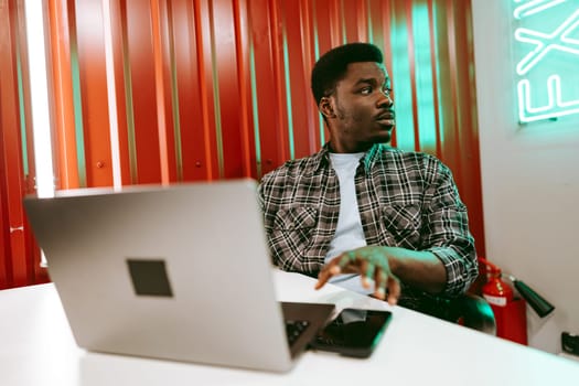 A young man sits at a white desk, focused on his laptop. The workspace features striking red walls and green neon lights, creating an energetic atmosphere for productivity.