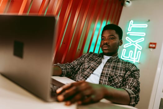 A young man focused on his laptop in a trendy workspace. The red wall features a glowing exit sign, creating a vibrant atmosphere during the daytime.