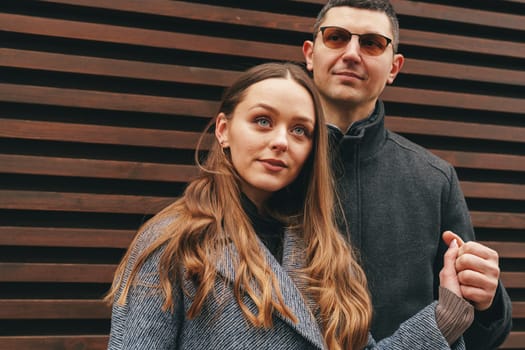 A couple stands closely together, smiling and holding hands, in front of a modern wooden wall. Their casual attire complements the relaxed atmosphere of the sunny day.