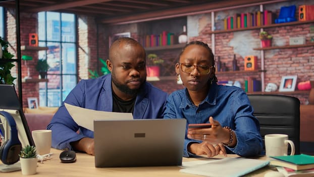 Mature couple dealing with online banking together at home in their loft, setting up payments and confirming money transfer. Pleased man and woman enjoy online shopping on laptop. Camera A.