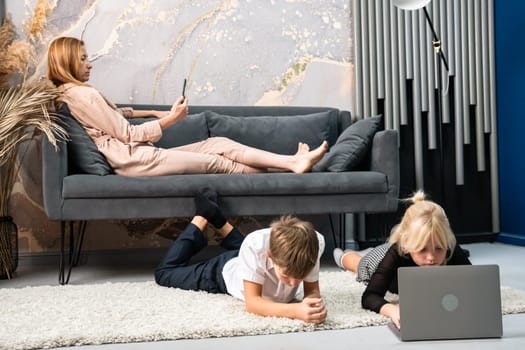A woman in a pink suit relaxes on a couch while two children lay on the floor, one using a laptop and the other playing with a handheld device.