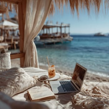 A serene beachfront scene with a laptop, an open book, a refreshing drink, and cozy cushions, offering a perfect setting for remote work or relaxation.
