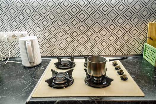 Close-up of a built-in gas stove in a kitchen interior, with a small saucepan. The gas hob is light beige, next to it is a white electric kettle.