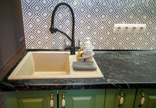 Detail of a light beige kitchen sink with a modern black faucet, dark green marble countertop, and a black and white tile backsplash. Dishwashing accessories.