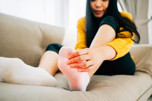 A woman seated on a sofa massages her painful foot highlighting the area of discomfort. Illustrating health care reflexology and recovery through massage and relief from pain.