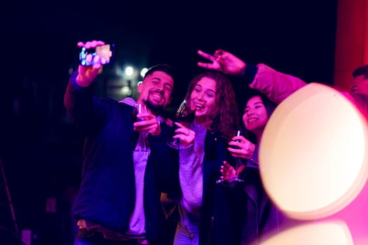Four friends gather together at nighttime, enjoying drinks and taking selfies. The atmosphere is festive with colorful lights enhancing the joyful mood of the celebration.