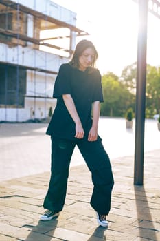 A young woman stands on a cobblestone path in a casual black outfit, surrounded by an urban environment under soft evening light, showcasing her relaxed style.