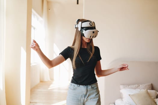 A woman in a casual black t-shirt and jeans is immersed in a virtual reality activity at home. She appears to be interacting with digital content in a bright, sunlit room.