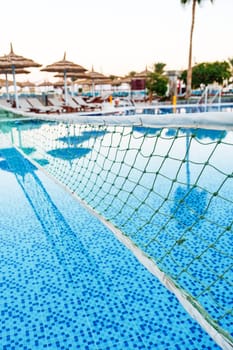 A tranquil swimming pool glistens under the warm sunset, featuring a volleyball net stretched across the water. Lounge chairs and palm trees create a relaxing atmosphere.