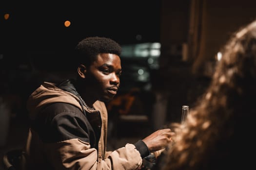 Under the dim lights of a night gathering, a young man listens intently while seated at an outdoor cafe, with the city lights softly illuminating the background.