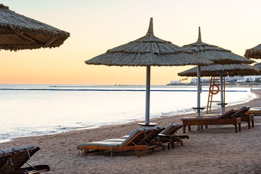 Sunlit beach features thatched umbrellas and lounge chairs arranged neatly, offering a serene spot for relaxation by the ocean during sunset glow.
