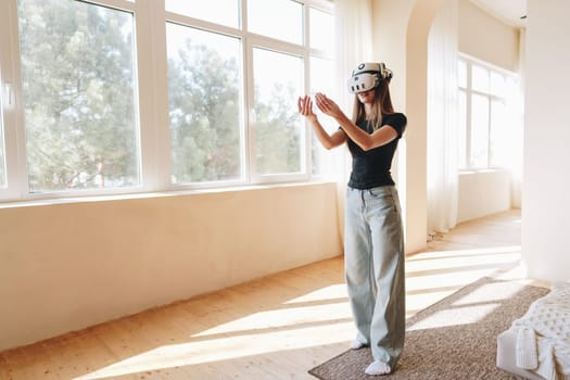 A woman is fully immersed in a virtual reality session, wearing a headset and using hand gestures to interact. The spacious room is filled with natural light, highlighting her concentration.