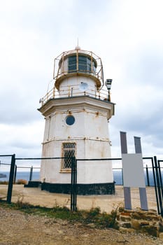 A weathered lighthouse sits atop a rocky terrain, surrounded by a fence. The cloudy sky looms overhead, and lookout equipment is mounted for navigation.
